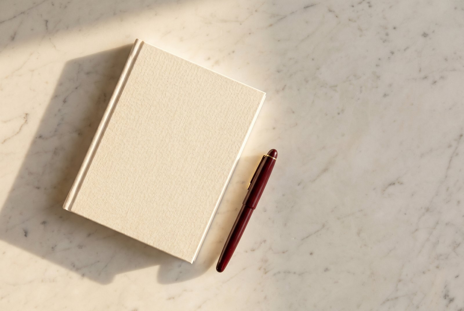 Closed cream notebook and oxblood fountain pen on cream marble, late-morning side light.